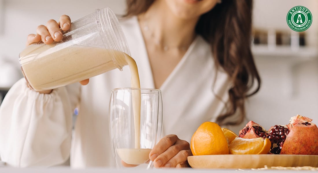 Woman pouring a creamy detox drink into a glass beside a plate of fresh fruit